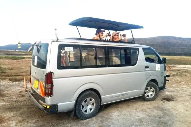 Tourists in a pop-up roof safari vehicle photographing wildlife in Nairobi National Park with the city visible in the background